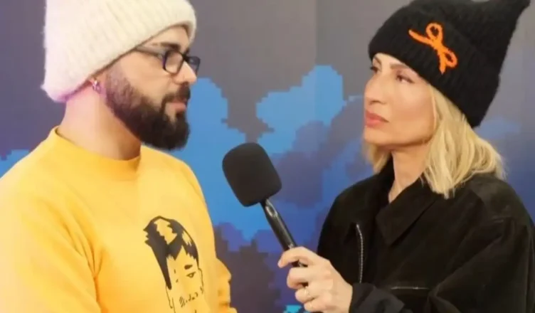 Ο τραγουδιστής Ακύλας φοράει το χαρακτηριστικό του σκουφάκι και χαμογελά κατά τη διάρκεια συνέντευξης για τη Eurovision 2026.
